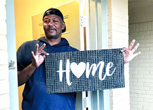A HUD resident stands in his doorway, holding up a sign that says ‘HOME’ with a heart in it.